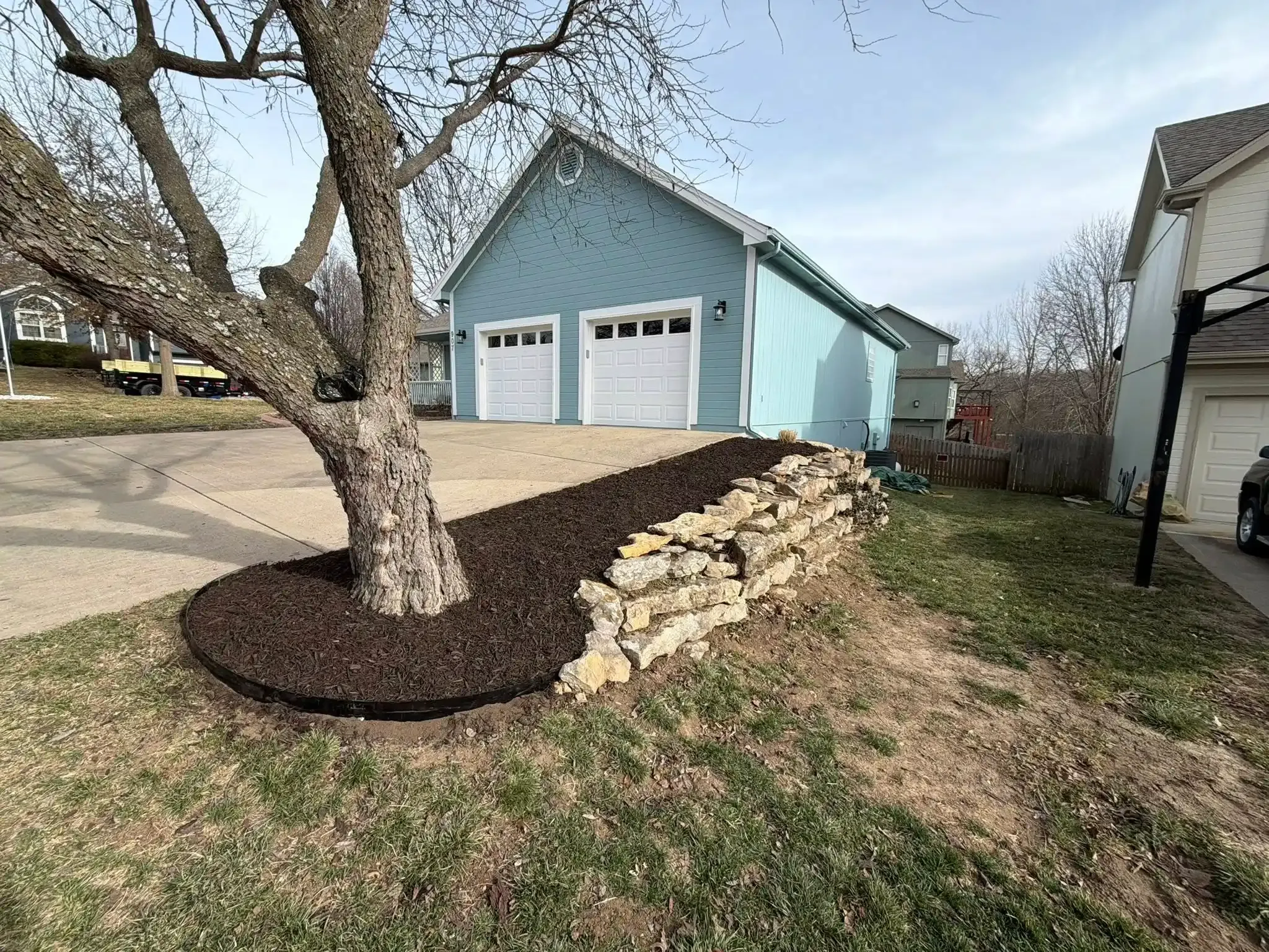 Natural fieldstone retaining wall with mulch bed and tree ring along driveway — landscaping and retaining walls in Smithville MO